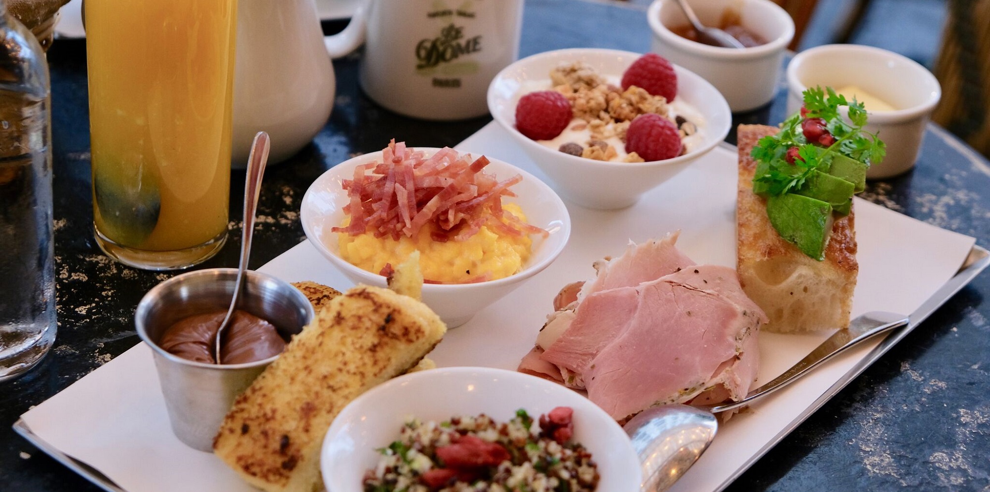 Table de brunch en terrasse avec boissons, tartines d'avocat, croissants et ambiance parisienne